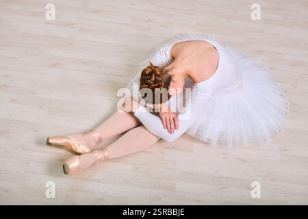 Giovane ballerina femminile con capelli auburn in tutù bianco si siede su un pavimento di legno chiaro, la testa poggia sulle ginocchia. La sua posa e la sua espressione trasmettono tristezza ed ex Foto Stock