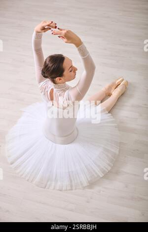 Giovane ballerina femminile con capelli scuri in tutù bianco classico seduta su pavimento in legno, braccia sollevate elegantemente. L'illuminazione soffusa mette in risalto la postura aggraziata Foto Stock