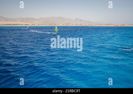 Il luminoso pomeriggio cattura i windsurfer che scivolano attraverso le vivaci acque blu del Mar Rosso. Le montagne costiere formano uno sfondo lontano sotto il cielo limpido. Prospettiva Foto Stock