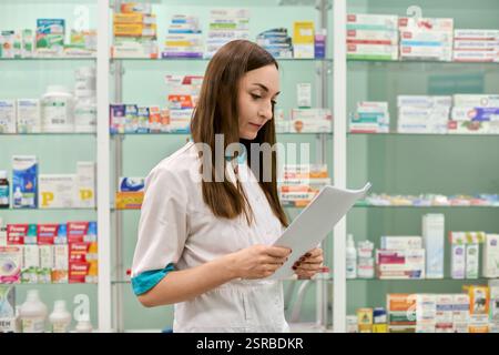 Giovane donna farmacista adulta con capelli castani, caucasico, revisione prescrizione in farmacia illuminata. Scaffali di medicinali e prodotti in backgr Foto Stock