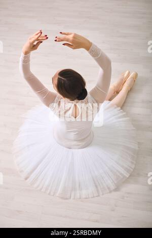 Giovane ballerina femminile con capelli scuri in tutù bianco classico seduta su pavimento in legno, braccia sollevate elegantemente. L'illuminazione soffusa mette in risalto la postura aggraziata Foto Stock