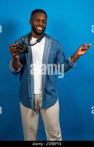 Giovane fotografo che tiene in mano una fotocamera professionale e punta da parte con l'indice, sorridendo su sfondo blu Foto Stock