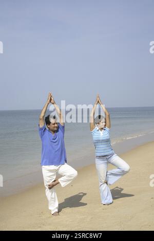 Giovane uomo e donna dell'India meridionale che fa yoga in piedi su una gamba piegata e solleva palme articolari sopra la riva del mare, Shiroda, Dist. Sindhu Foto Stock