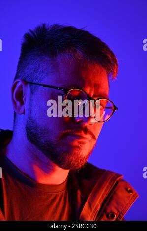Foto elegante di un giovane con occhiali ottici e piccola barba di classe con illuminazione blu-rosa su sfondo viola con luce al neon. Bellezza, arte, moda Foto Stock