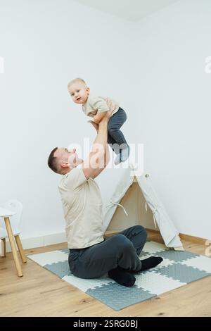 Un padre gioioso solleva il suo bambino in alto in una stanza luminosa e allegra dotata di tenda da gioco Foto Stock