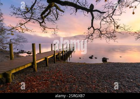 Il sole di Derwentwater sorge in una mattina d'autunno nebbiosa con un vecchio molo di legno che conduce all'esterno su acque calme. Lake District, Regno Unito. Foto Stock