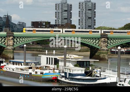 Londra, Regno Unito. Un treno della London Overground attraversa l'Hammersmith Bridge sul Tamigi, con case galleggianti ormeggiate sotto. Foto Stock