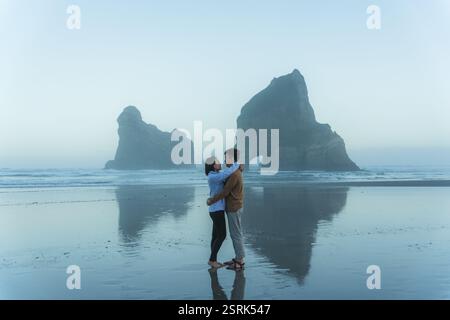 Una romantica coppia asiatica condivide un momento tenero sulla spiaggia di Wharariki con le isole Archway nel mare di Tasman a Cape Farewell, nell'Isola del Sud della nuova Zelanda Foto Stock