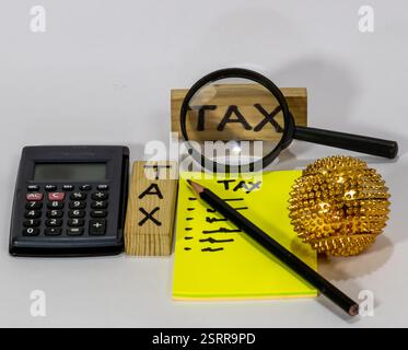 C'è una calcolatrice nera con bottoni rossi e bianchi, una lente d'ingrandimento, una pila di monete e un blocco di legno con la parola "TAX" scritta sopra. TH Foto Stock