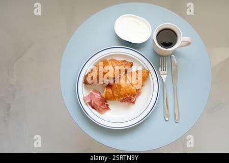 Due croissant classici con prosciutto e formaggio spalmabile con caffè nero sulla vista del tavolo blu Foto Stock