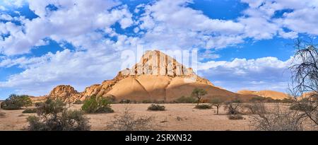 Piccola montagna di Spizkoppe sotto nuvole bianche che coprono il cielo blu, cespugli e un paio di cottage, Spitzkoppe, Namibia, Africa Foto Stock