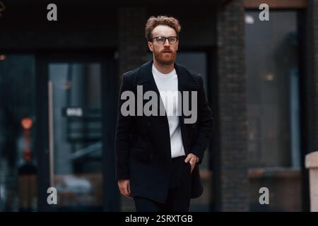 Un uomo elegante con barba e occhiali si trova all'aperto vicino all'edificio. Foto Stock