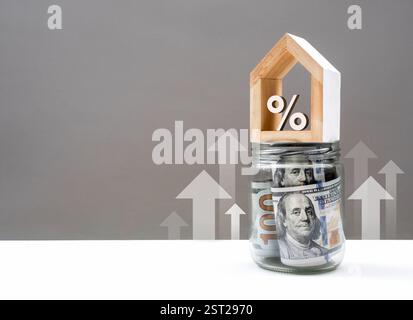 Icona percentuale 3d in casa bianca su vaso di vetro con banconote da cento dollari, con frecce in aumento su tavolo bianco, sfondo grigio. Risparmio mese Foto Stock