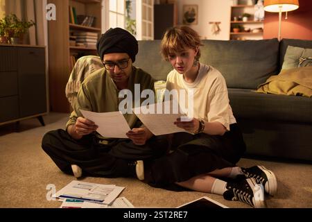 Due giovani adulti seduti sul pavimento, concentrati sulla revisione di bollette e documenti finanziari, dimostrando collaborazione e discussione in un accogliente salotto Foto Stock