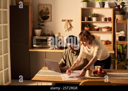 Due colleghi che lavorano al bancone della cucina, discutono documenti e idee in un ambiente accogliente e moderno con piante e scaffali pieni di vari oggetti Foto Stock