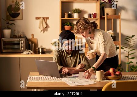 Giovane coppia che lavora insieme a casa su un progetto utilizzando laptop e documenti su un tavolo da cucina. Luce seminale che illumina la cucina moderna con varie piante della casa è visibile Foto Stock