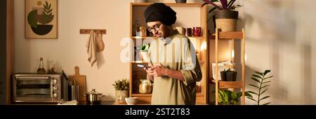 Uomo con turbante sorridente mentre controlla il telefono in una cucina ben illuminata con piante e scaffali in legno Foto Stock