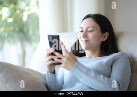 Donna asiatica che si rilassa controllando lo smartphone seduto su un divano a casa Foto Stock