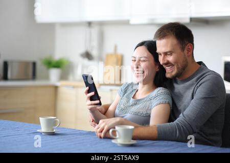 Felice coppia interrazziale che ride controllando lo smartphone in cucina a casa Foto Stock