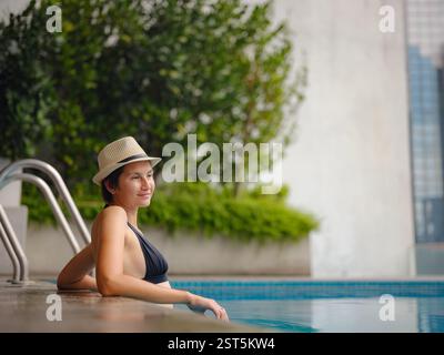 Giovane donna asiatica che si rilassa a bordo piscina all'hotel Kuala Lumpur con vista sui grattacieli circostanti, godendo del tempo libero in un vivace ambiente urbano. Foto Stock