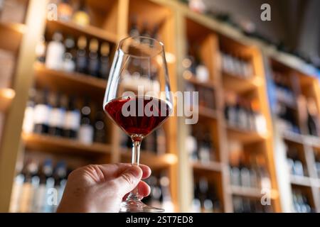 Una mano di mans tiene in primo piano un buon bicchiere di vino di cristallo con una cantina toscana su grandi scaffali in legno non focalizzato. Concetto di degustazione Foto Stock