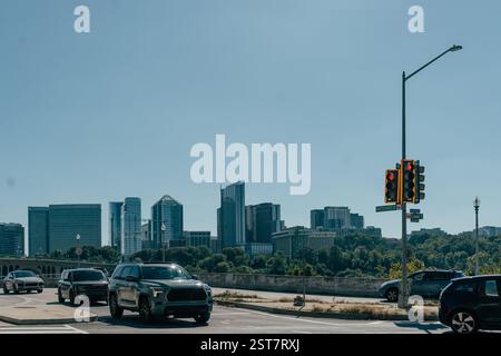 Le auto sono fermate a un semaforo vicino a una sponda del fiume, che mostra un vivace skyline della città con architettura moderna e verde sullo sfondo su una S. Foto Stock