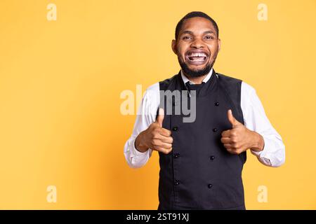 Maggiordomo felice e sicuro che dà un simbolo di pollice davanti alla telecamera, presentando il suo accordo e le emozioni positive in studio. Giovane cameriere che mostra un segno simile. Foto Stock