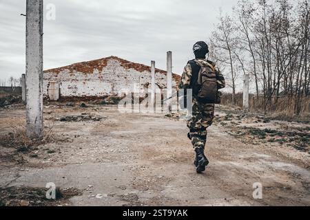Foto di un soldato completamente equipaggiato con giubbotto corazzato, casco, occhiali per il viso e protezione che attacca con la pistola sullo sfondo della città distrutta. Foto Stock