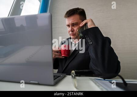 Il giovane uomo d'affari in ufficio comunica con uno smartphone e beve il tè da una tazza. Flusso di lavoro quotidiano in ufficio. Foto Stock