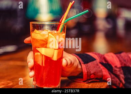 Uomo che regge un bicchiere di cocktail negroni. Mano maschile con bicchiere di cocktail Aperol spritz. Cocktail con tubo su sfondo luminoso Foto Stock