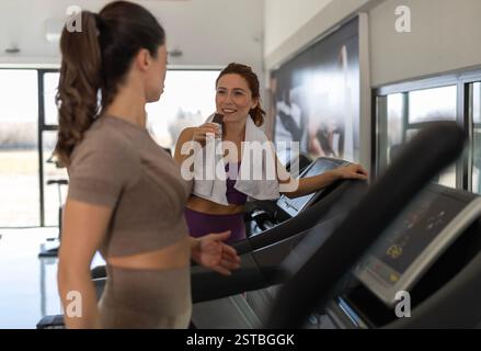 Due donne si impegnano in una conversazione mentre utilizzano tapis roulant, mentre si allenano in una palestra moderna durante il giorno Foto Stock