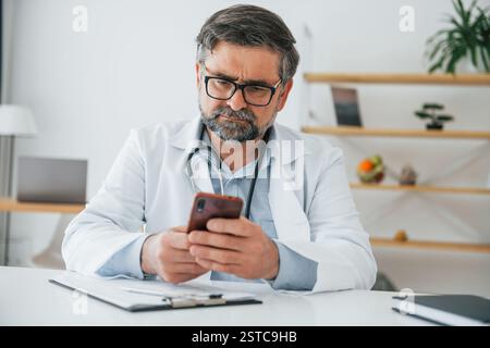 Un medico maschile è seduto con il telefono in mano. Operatore medico professionale in cappotto bianco è in ufficio. Foto Stock