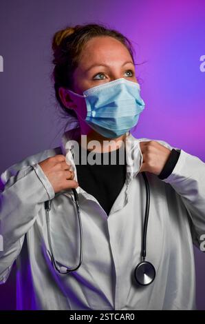 Giovane donna medico professionista che indossa una maschera e uno stetoscopio, regolando il suo camice da laboratorio in un ambiente colorato e moderno Foto Stock