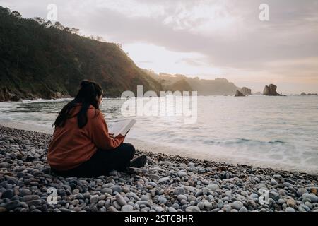 Una donna con cappuccio arancione e pantaloni neri si siede su una spiaggia di ciottoli a Playa del Silencio, Asturie, Spagna, leggendo un libro mentre guardi il tramonto Foto Stock