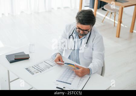 L'uomo sta lavorando con i documenti. Un medico maturo in giacca bianca è in ufficio. Foto Stock