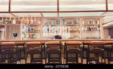 Un moderno interno bar con bottiglie di alcol e una barista che lavora dietro il bar, senza clienti presenti Foto Stock