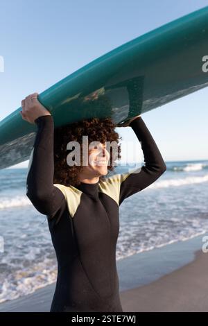 Vista laterale della giovane ragazza felice in muta con tavola da surf in piedi tenendo la tavola da surf sopra la testa guardando via sulla riva bagnata dal mare ondeggiante Foto Stock