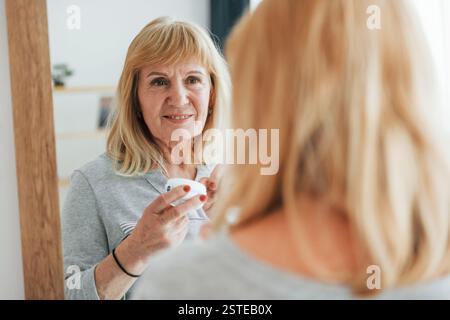 Guarda lo specchio, cura di sé. Una donna anziana con i capelli rossi è a casa da sola. Foto Stock
