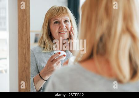 Guarda lo specchio, cura di sé. Una donna anziana con i capelli rossi è a casa da sola. Foto Stock