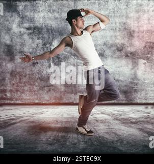 Un giovane uomo con una canotta e un cappello che fa una routine di danza in posa su una gamba in una sala o un palco in cemento grunge Foto Stock