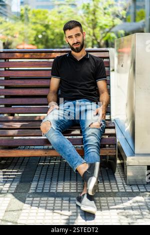 Un giovane uomo alla moda con barba e jeans alla moda si siede casualmente su una panchina di legno in un parco illuminato dal sole, trasudando un senso di rilassato stile di vita urbano Foto Stock