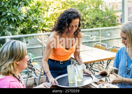Un trio di amiche di mezza età si gode un incontro informale su una terrazza soleggiata all'aperto, condividendo risate e bevande Foto Stock