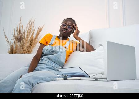 La donna afroamericana soffre di mal di testa mentre lavora su un laptop sul divano. Elegante ragazza freelance nera che lavora a casa online Foto Stock