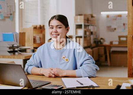 Donna sorridente seduta alla scrivania in un ufficio moderno, con penna, computer e documenti sulla scrivania, circondata da forniture e arredi moderni Foto Stock