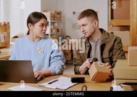 Colleghi che collaborano in un moderno ufficio di magazzino, donna ispanica e uomo caucasico discutono di logistica mentre utilizzano la tecnologia, circondati da pacchi e documenti Foto Stock