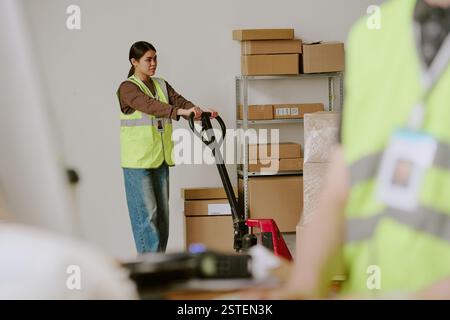 Giovane donna che lavora in magazzino, sposta scatole con martinetto a pallet con giubbotto di sicurezza riflettente e si concentra sull'attività da svolgere Foto Stock