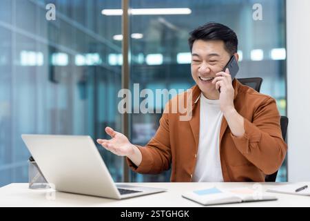 Uomo asiatico gioioso di successo che lavora all'interno dell'ufficio sul posto di lavoro. Uomo d'affari che parla al telefono cellulare seduto alla scrivania e che lavora con un computer portatile. Foto Stock