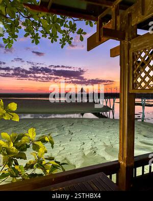 Tramonto su un Molo di legno Una vista da una terrazza di legno che si affaccia su una spiaggia con bassa marea, con un molo che si estende verso un padiglione distante. Il cielo brilla Foto Stock