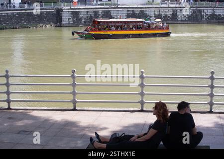 Una coppia di viaggiatori si rilassa riposando all'ombra lungo il fiume Singapore. Bumboat sullo sfondo traghetto per turisti. Fascino e paesaggio tranquillo con vista sulla città. Foto Stock