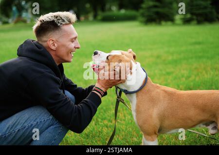 Il proprietario tiene un cane con una palla in bocca per la museruola. Un uomo caucasico con un sorriso gioca con una palla con il suo cane nel parco. Addestramento per cani. Ameri Foto Stock